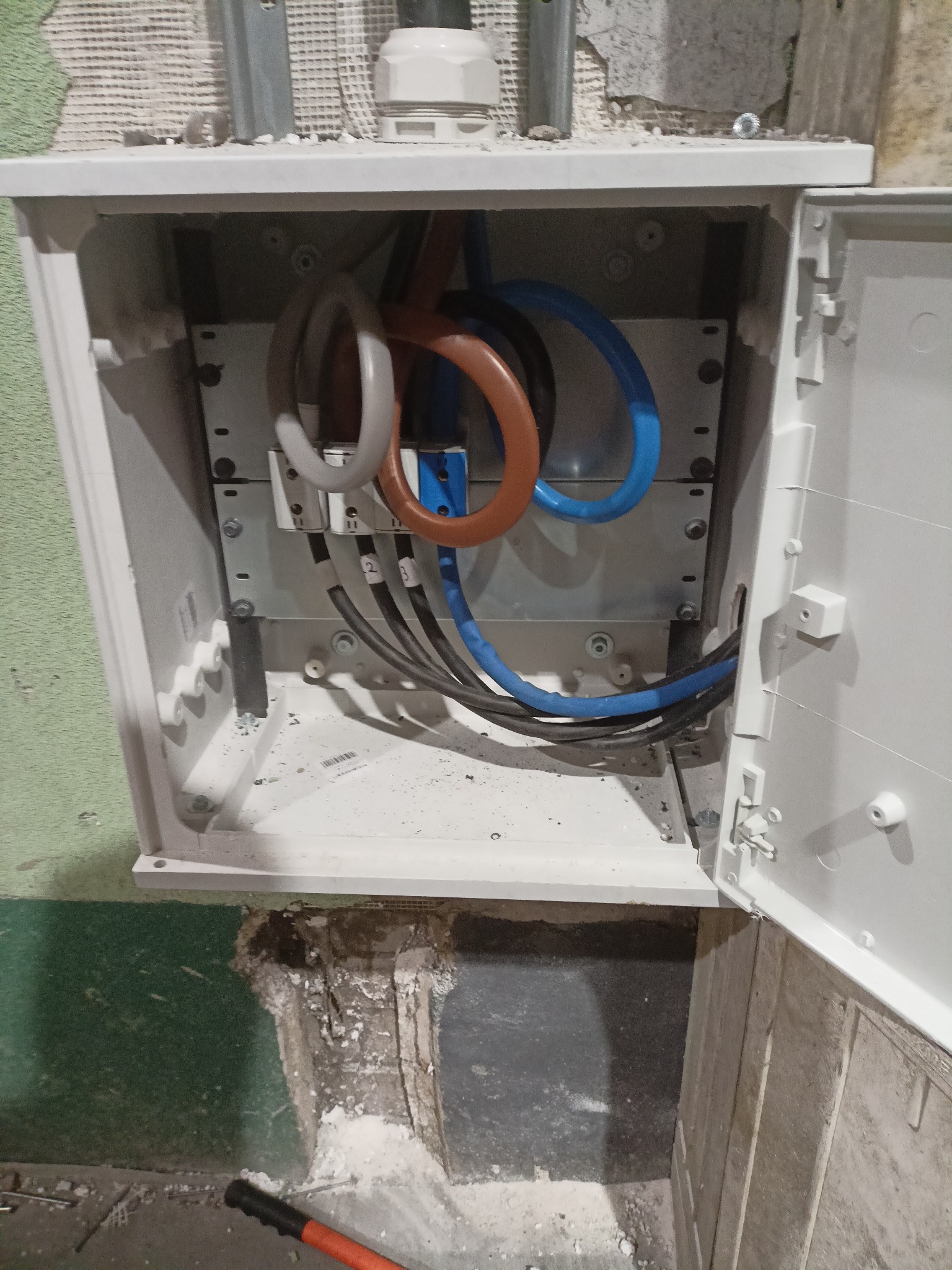 Otwarta, biała skrzynka elektryczna z kolorowymi przewodami wewnątrz, zamontowana na ścianie. Widoczne oznaczenia na kablach i fragment ściany z instalacją.