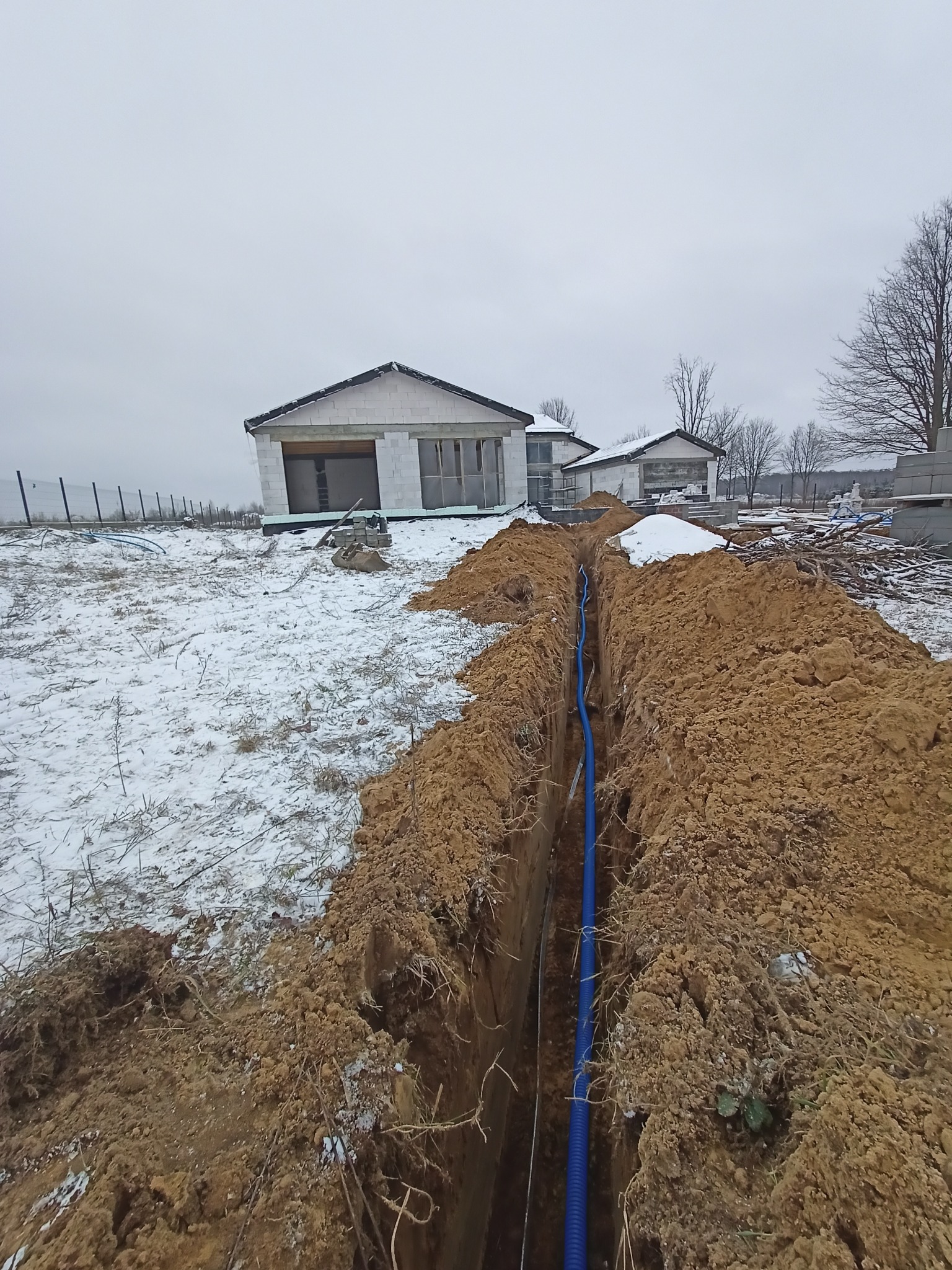 Wykop z niebieską rurą kablową na tle budowy domu w zimowej scenerii. Widok z góry na wykopaną ziemię, rurę i budynek w stanie surowym otwartym.