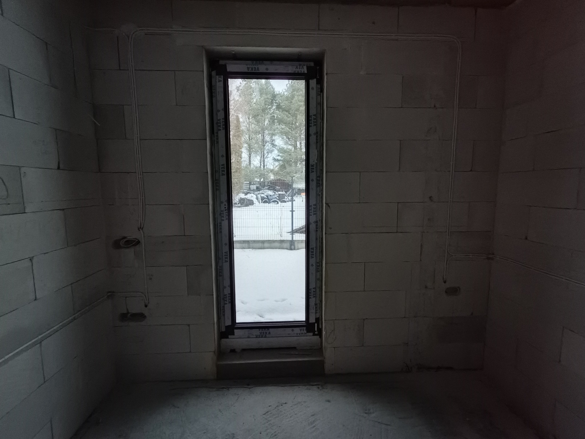 Surowe wnętrze z oknem na zimowy krajobraz. Widoczne kable elektryczne na ścianach z betonu komórkowego, sugerujące etap instalacji elektrycznej.