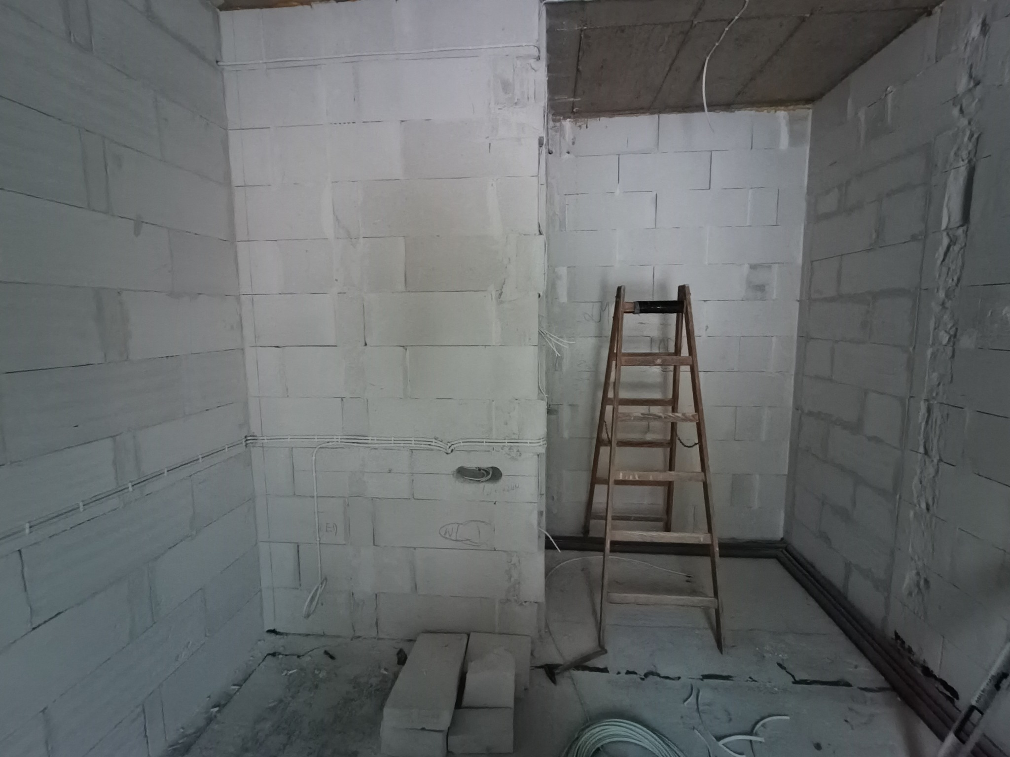 Surowy pokój z białych bloczków, widoczne kable elektryczne na ścianach i drabina. Instalacja elektryczna w trakcie realizacji, przygotowanie do dalszych prac wykończeniowych.
