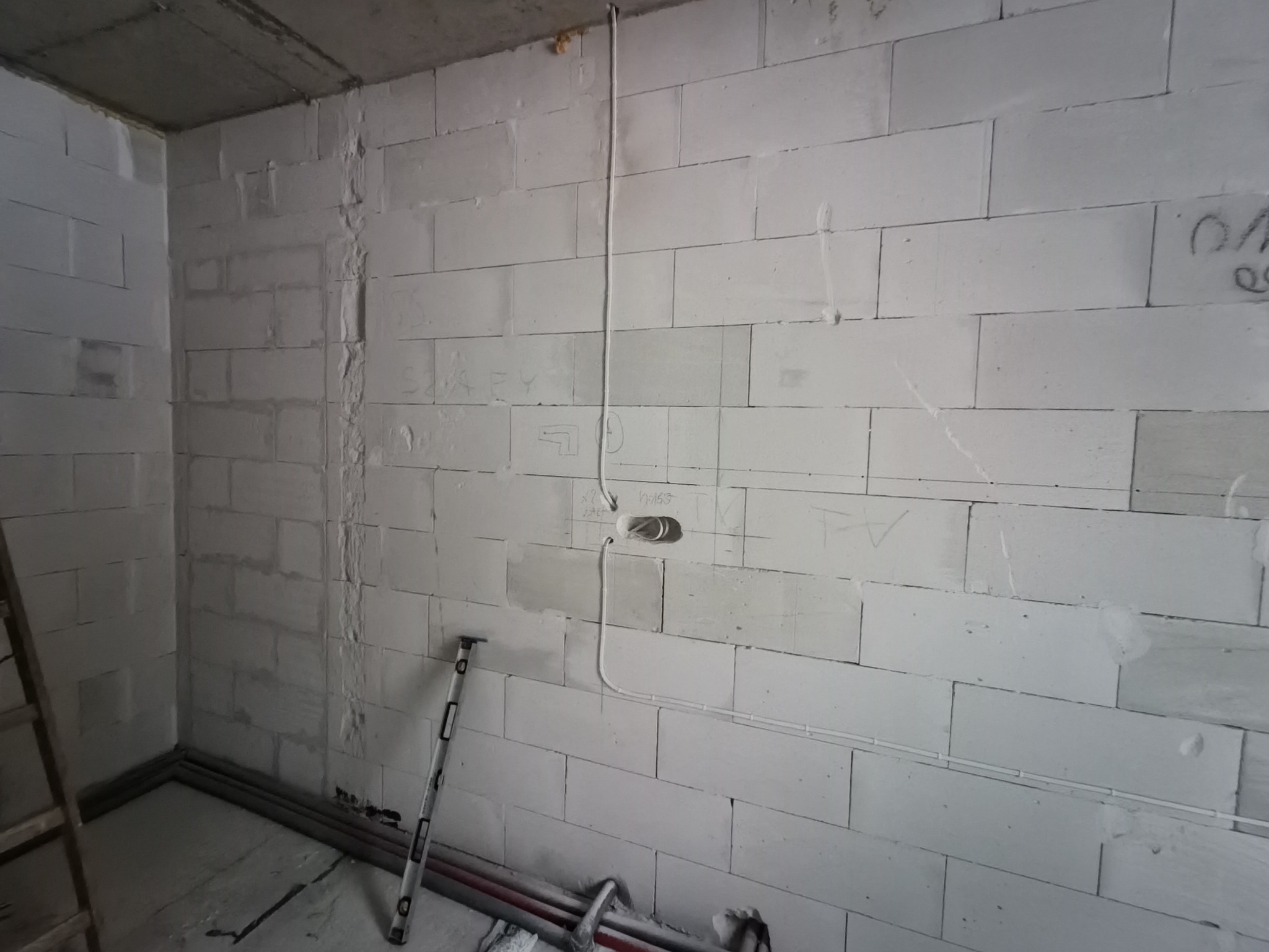 Wnętrze w trakcie prac elektrycznych: przewody prowadzone po ścianie z betonu komórkowego, poziomica oparta o ścianę, widoczne rury instalacyjne przy podłodze.