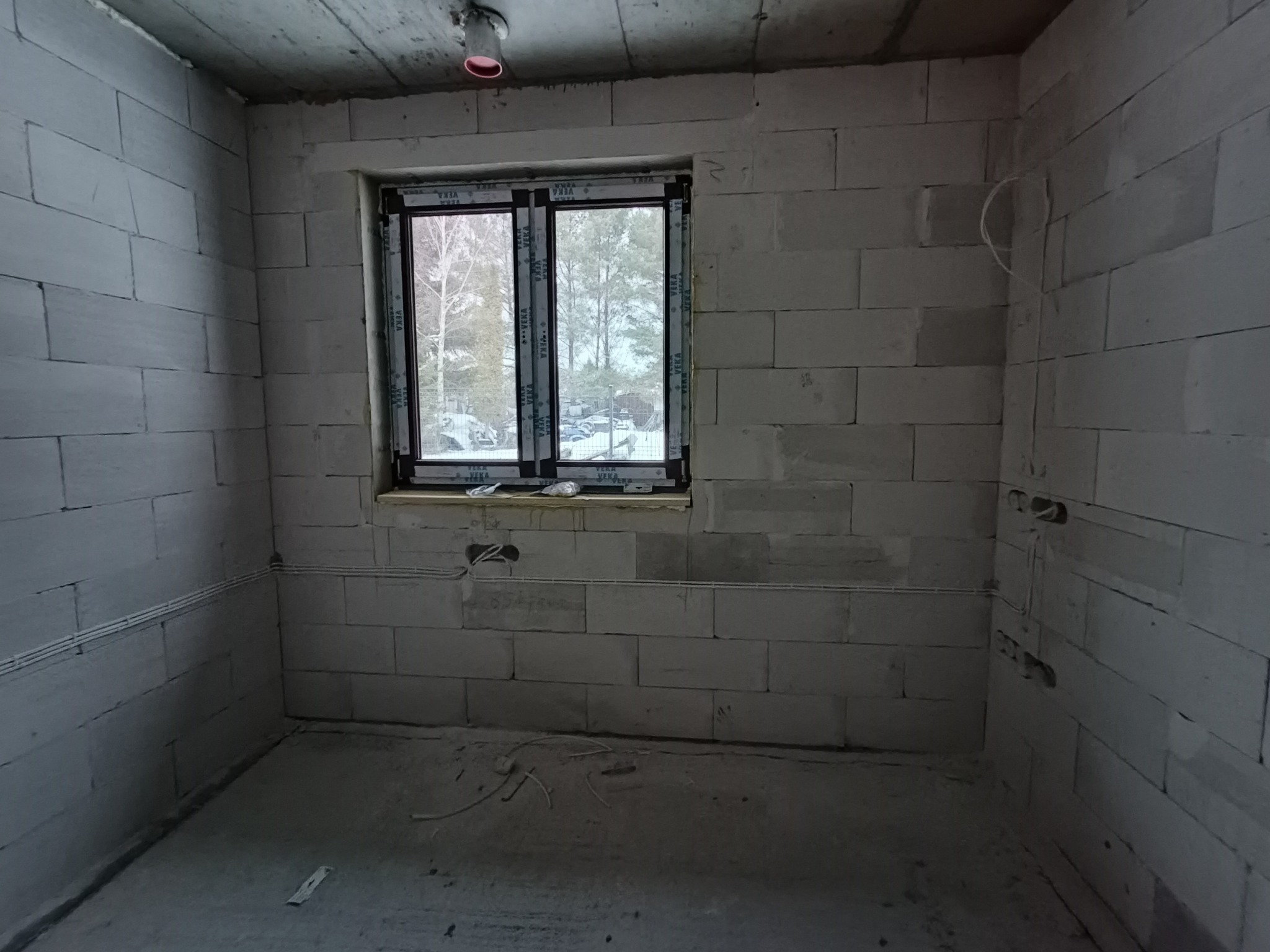 Wnętrze surowego pomieszczenia z oknem, widoczne przewody elektryczne w ścianach z betonu komórkowego. Instalacja w trakcie realizacji, przygotowanie do wykończenia.