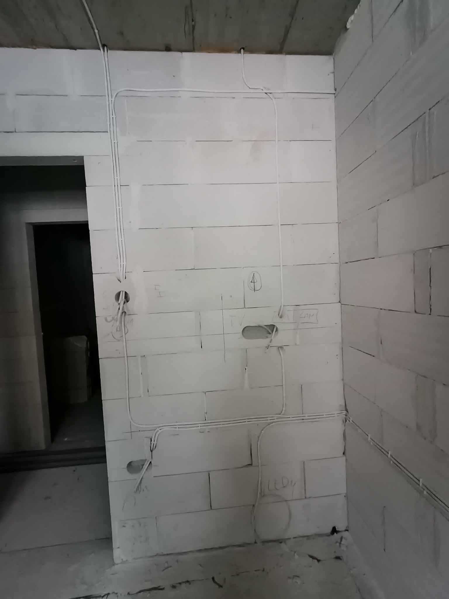 Surowe ściany z betonu komórkowego z rozprowadzoną instalacją elektryczną, widoczne kable i puszki pod gniazdka, oznaczenia na ścianie. Etap prac instalacyjnych.