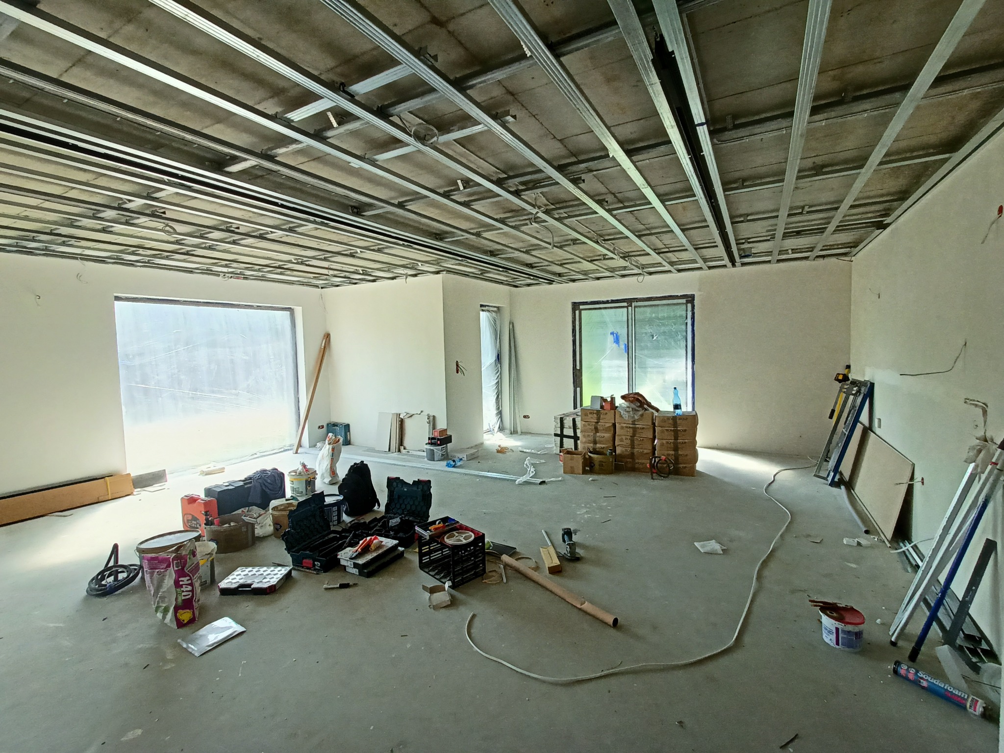 Wnętrze w trakcie prac: widoczne metalowe profile sufitowe podwieszane, narzędzia, drabiny i materiały budowlane. Surowy stan, przygotowanie do dalszych prac wykończeniowych.