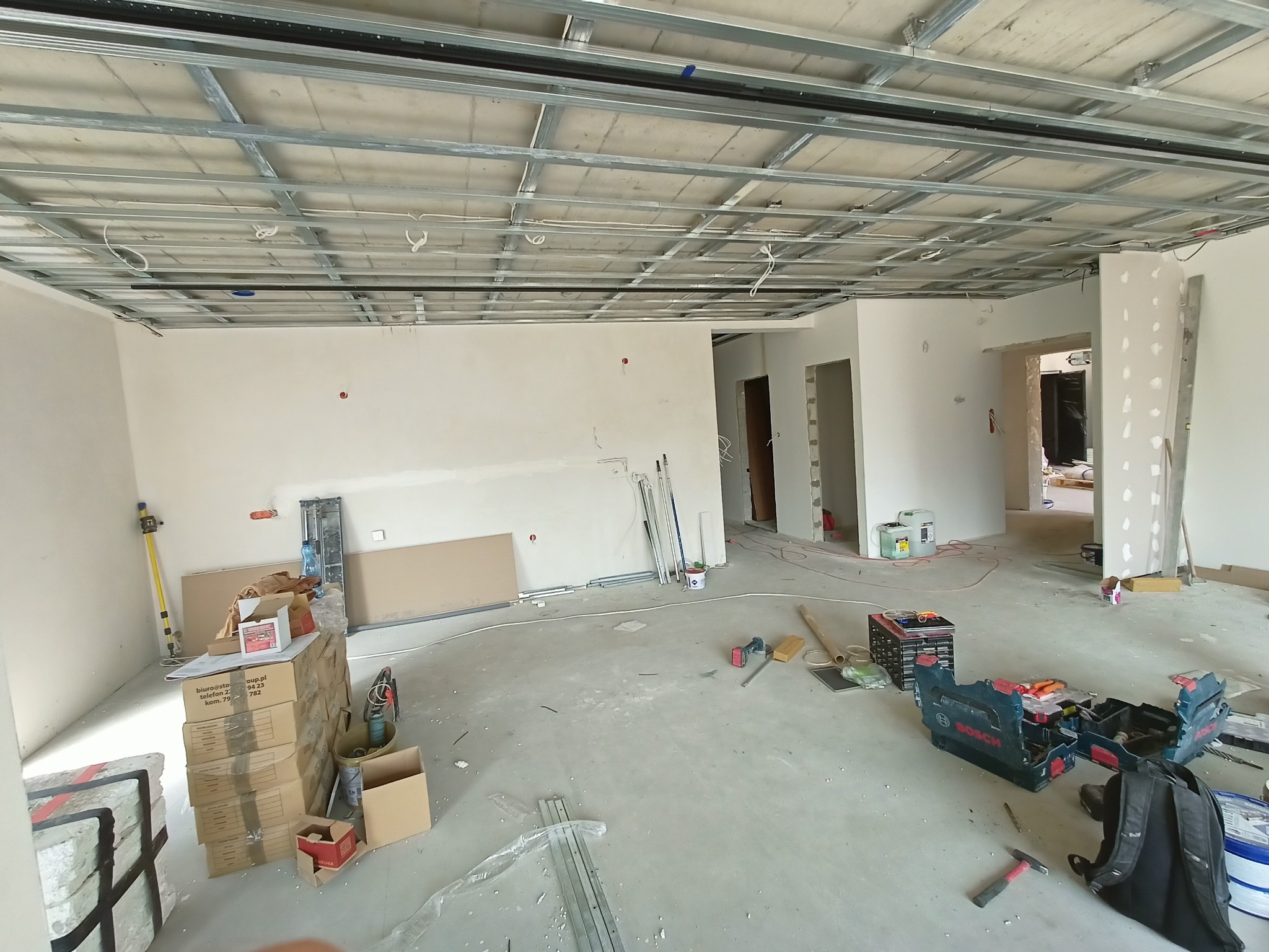 Wnętrze w trakcie remontu: widoczna metalowa konstrukcja sufitu podwieszanego, narzędzia i materiały budowlane. Stan surowy, przed wykończeniem.