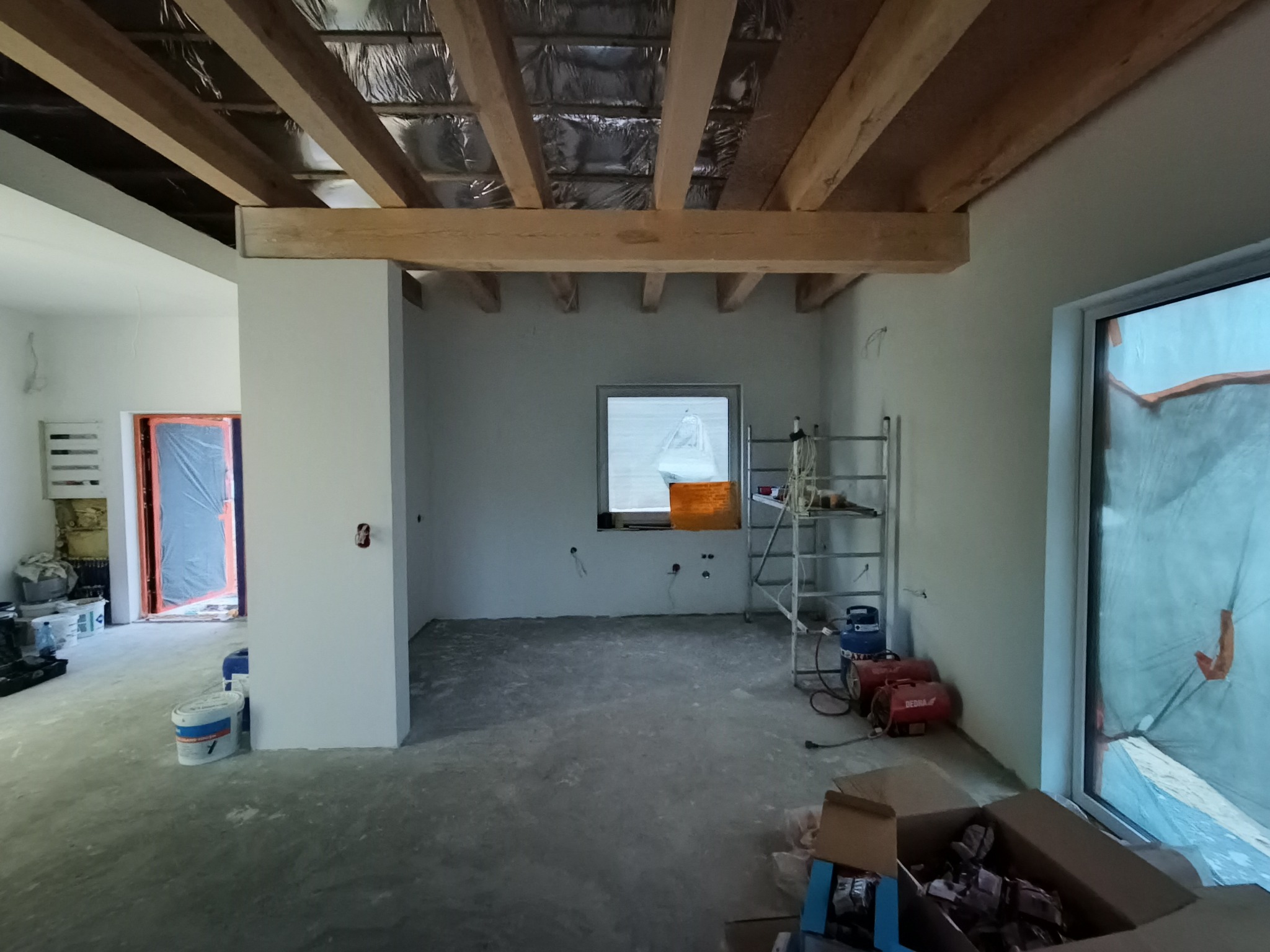 Wnętrze w trakcie prac wykończeniowych: surowy strop drewniany, ściany po gładzi, widoczne przewody elektryczne, drabina i nagrzewnice. Etap przed montażem oświetlenia.