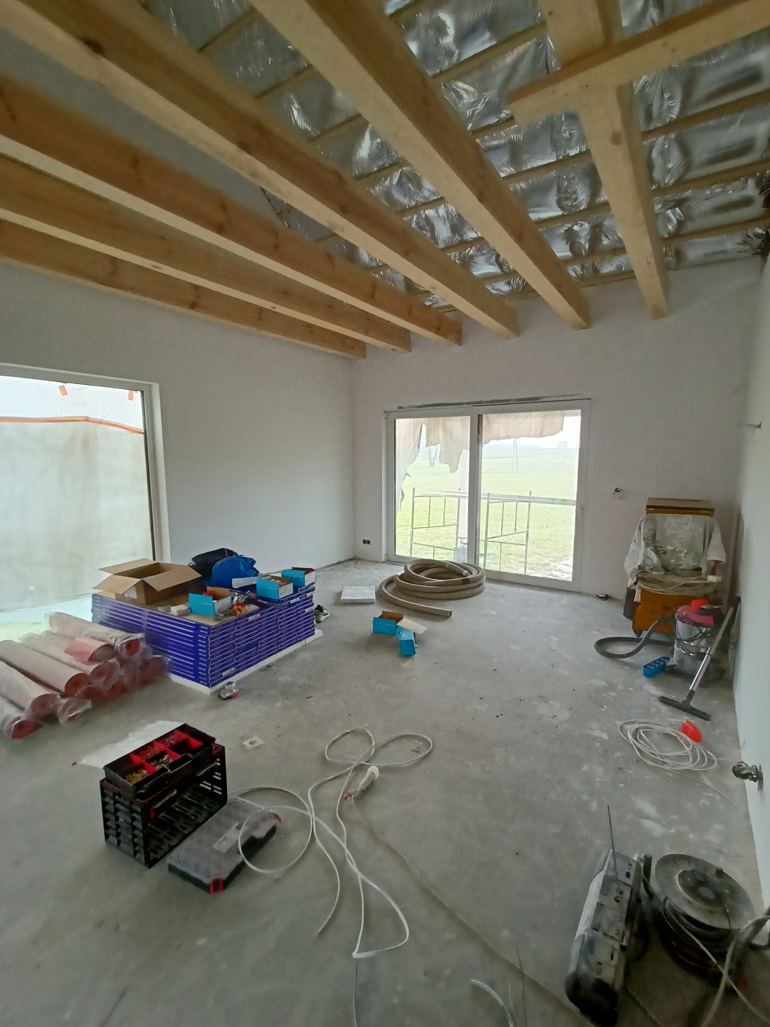 Wnętrze w stanie surowym z drewnianą konstrukcją dachu i narzędziami elektrycznymi. Widoczne kable, skrzynka z akcesoriami i rolki materiałów izolacyjnych.