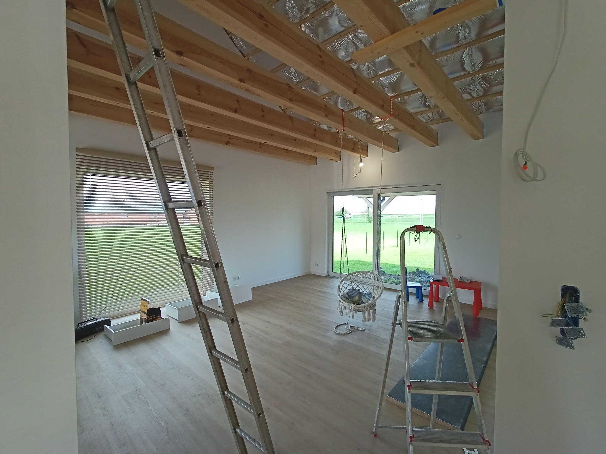 Wnętrze w trakcie prac wykończeniowych: surowy strop z izolacją, drabiny, widoczne kable elektryczne w ścianie. Przygotowanie do montażu instalacji elektrycznej.