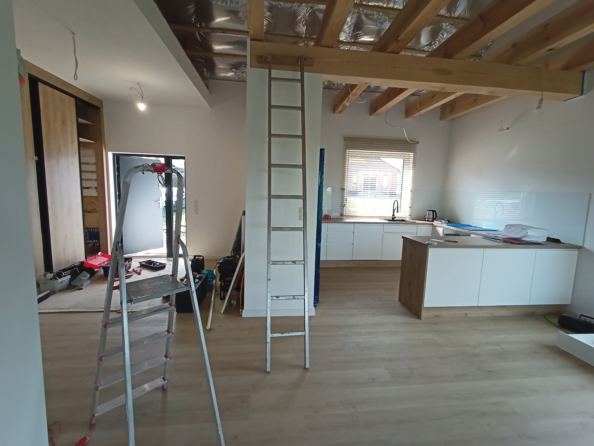 Wnętrze w trakcie prac wykończeniowych: widoczne drabiny, surowy sufit z izolacją, częściowo umeblowana kuchnia. Oświetlenie tymczasowe, podłoga wyłożona panelami.