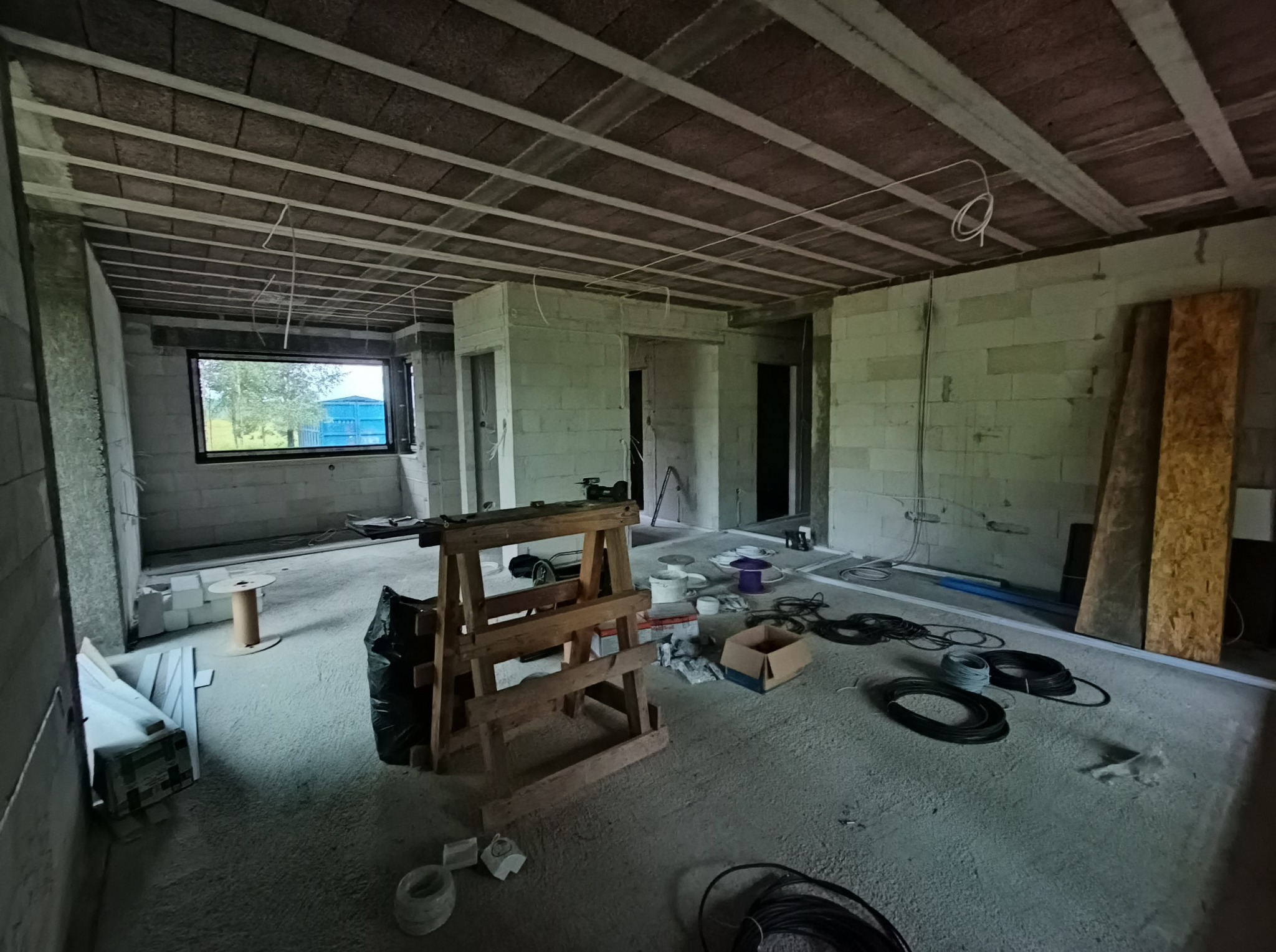 Surowy stan domu w budowie: widoczne przewody elektryczne zwisające z sufitu, kable na podłodze i drewniany kozioł. Okno z widokiem na zewnątrz.