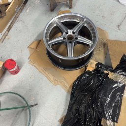 Szymon Gwóźdź - Aluminiowa felga samochodowa z pięcioramiennym wzorem, leżąca na kartonie w warsztacie. W tle piec i narzędzia, co sugeruje naprawę lub renowację.