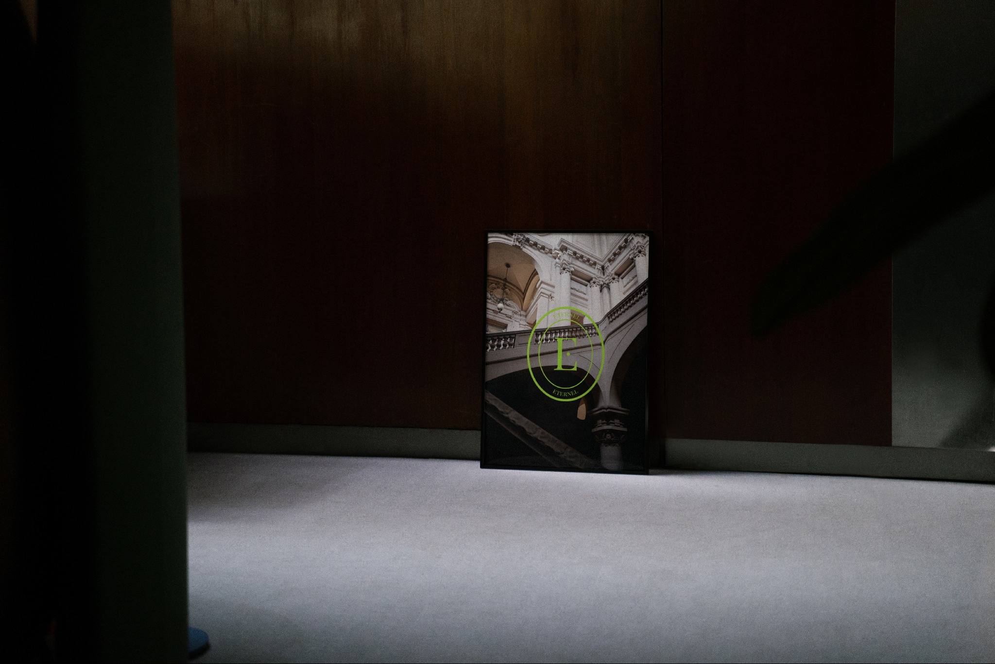 Plakat z wizualizacją wnętrza budynku z logo firmy, ustawiony na podłodze, w tle ściana. Prezentacja oferty agencji nieruchomości.