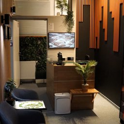PsycheClinic - Profesjonalna Psychoterapia i Wsparcie Psychologiczne Psycholog Warszawa Bemowo - Nowoczesne wnętrze poczekalni z recepcją, roślinami, regałem z książkami i dekoracyjnymi panelami ściennymi. Przytulny wystrój sprzyja relaksacji.