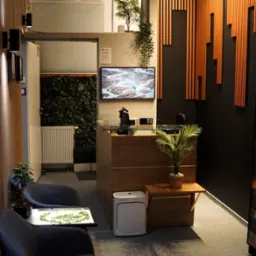 Nowoczesne wnętrze poczekalni z recepcją, roślinami, regałem z książkami i dekoracyjnymi panelami ściennymi. Przytulny wystrój sprzyja relaksacji.