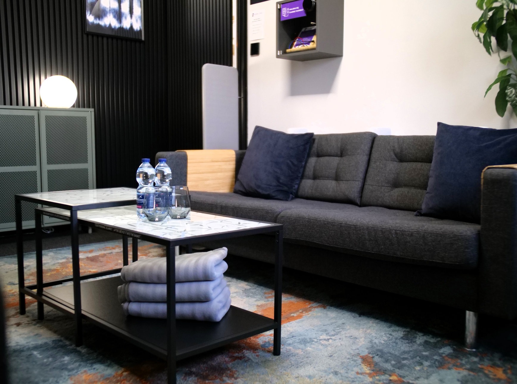 Wnętrze poczekalni: szara sofa z poduszkami, stolik z wodą i szklankami, dywan w odcieniach błękitu i pomarańczy, minimalistyczny design, lampa kulista.