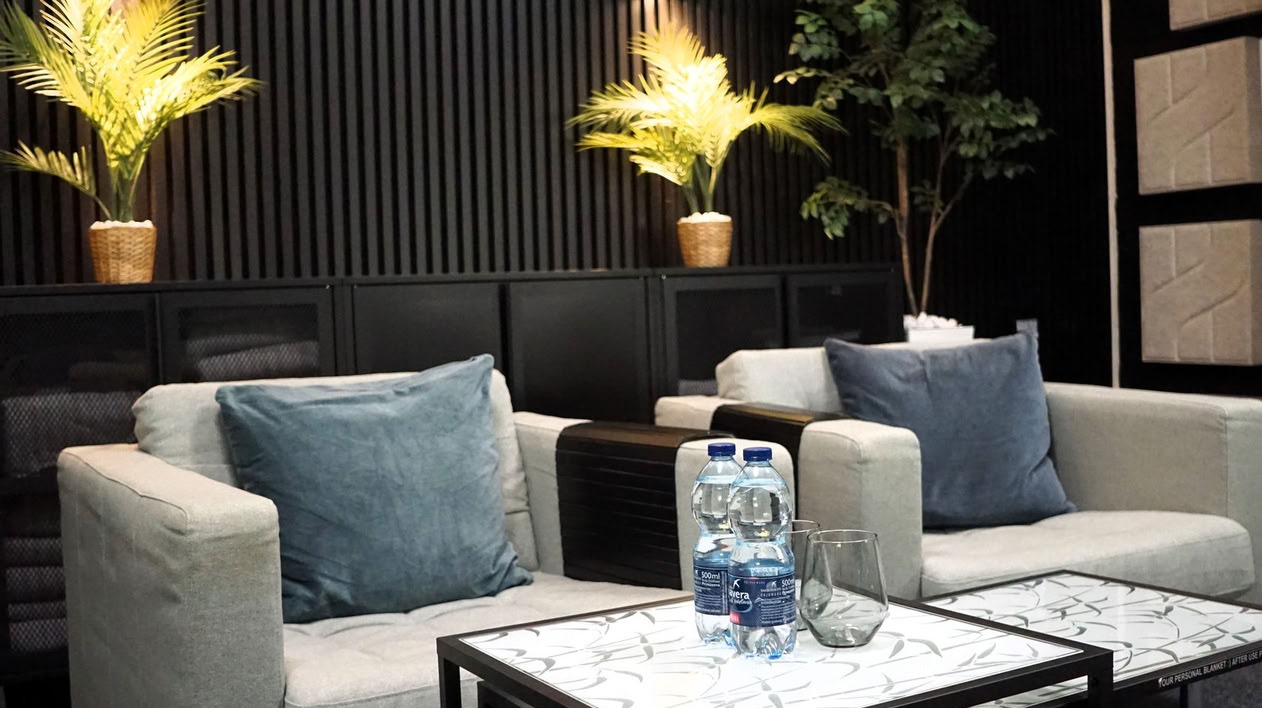 Wnętrze poczekalni z sofami, stolikami i roślinami doniczkowymi. Na stoliku woda i szklanki. Na ścianie panele akustyczne.