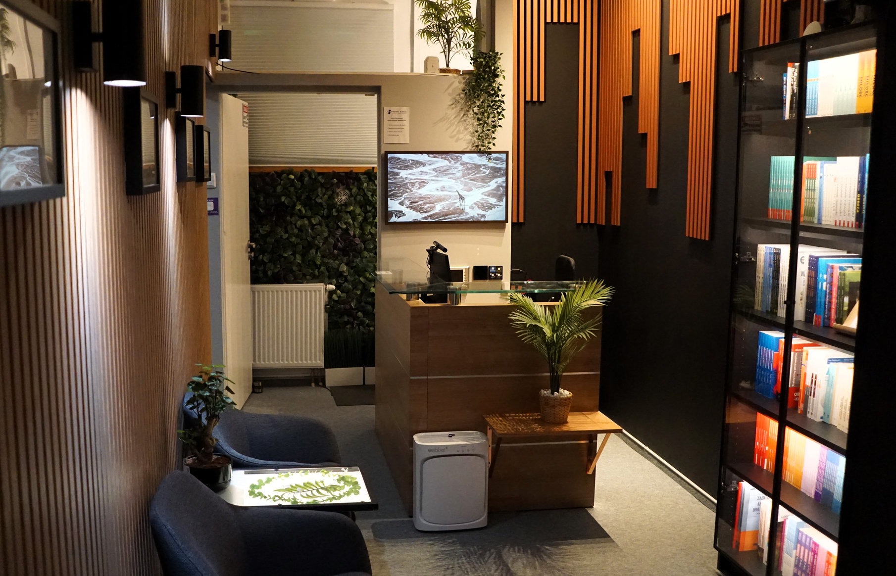 Nowoczesne wnętrze poczekalni z recepcją, roślinami, regałem z książkami i dekoracyjnymi panelami ściennymi. Przytulny wystrój sprzyja relaksacji.