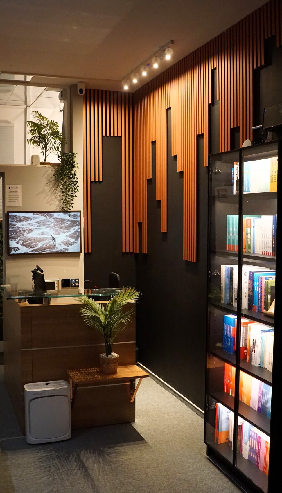 Recepcja w nowoczesnym gabinecie: drewniana lada, dekoracyjne lamele na ścianie, regał z książkami i rośliny doniczkowe. Minimalistyczny design.