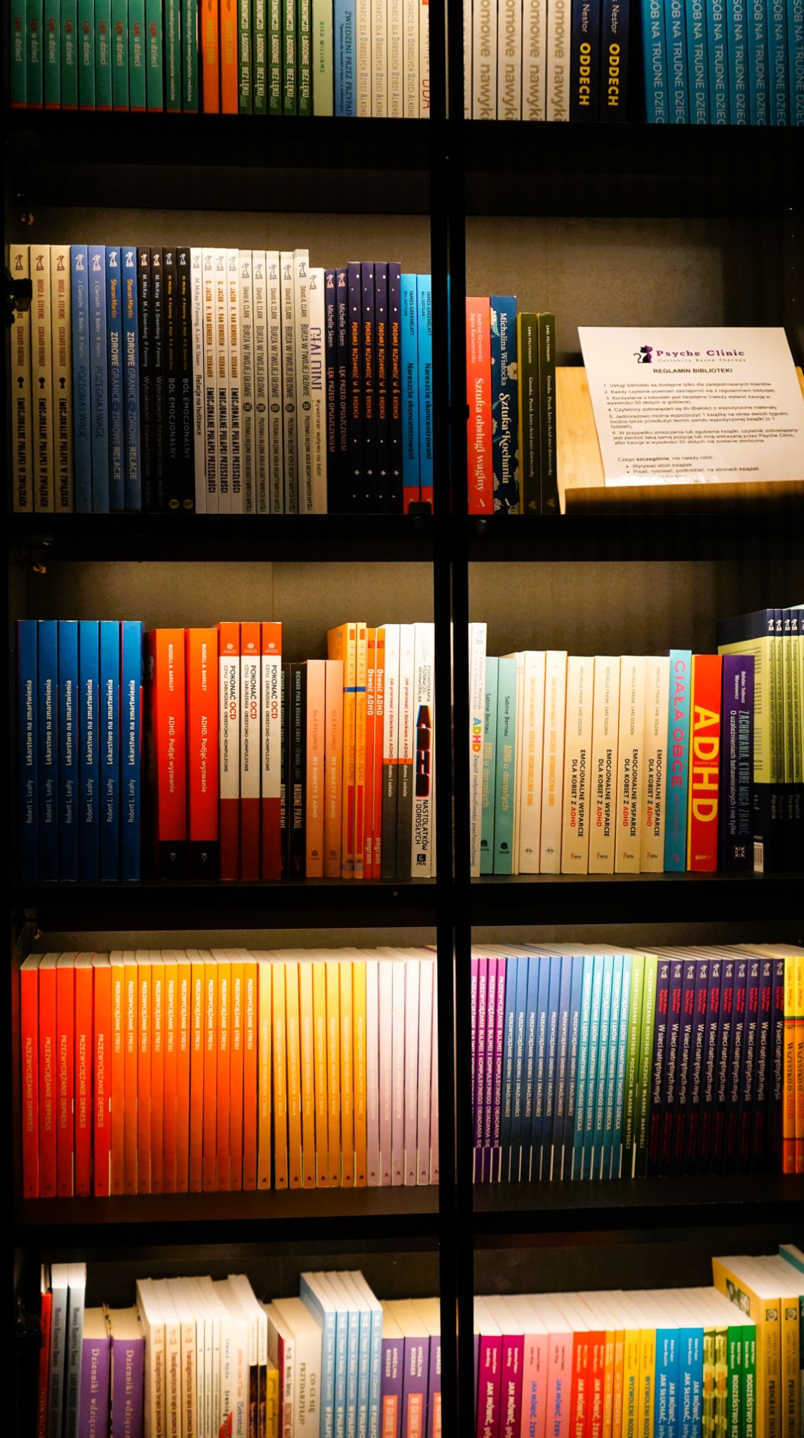 Półki z książkami w biblioteczce kliniki psychologicznej. Widoczne publikacje o ADHD, emocjach, relacjach, rozwoju osobistym. Na półce regulamin biblioteki.