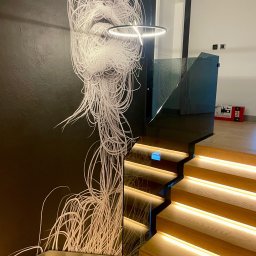 KM Glass - Nowoczesne schody z podświetleniem LED i szklaną balustradą. Na ścianie dekoracyjna grafika w stylu abstrakcyjnym. Minimalistyczne wnętrze z elementami drewna.