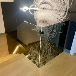 KM Glass - Nowoczesna, szklana balustrada schodowa z podświetleniem LED, odbijająca abstrakcyjny wzór na ścianie. Jasne, drewniane schody i minimalistyczne oświetlenie.