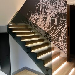KM Glass - Drewniane schody z podświetleniem LED i szklaną balustradą, wspornikowe, z abstrakcyjnym wzorem na ścianie. Nowoczesne wykończenie wnętrza, jasne drewno.