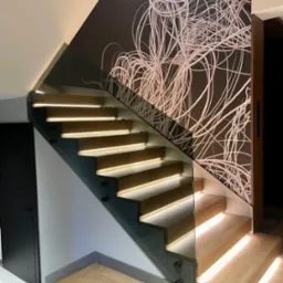 Drewniane schody z podświetleniem LED i szklaną balustradą, wspornikowe, z abstrakcyjnym wzorem na ścianie. Nowoczesne wykończenie wnętrza, jasne drewno.
