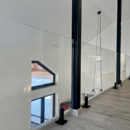 KM Glass - Nowoczesna balustrada ze szkła i czarnych słupków w jasnym wnętrzu. Widok z góry na drewnopodobną podłogę i okna z widokiem na zimowy krajobraz.