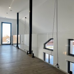 KM Glass - Nowoczesna balustrada szklana z czarnymi słupkami w jasnym wnętrzu domu. Widok na korytarz i okna. Podłoga z paneli imitujących drewno.