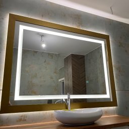 KM Glass - Łazienka z podświetlanym lustrem w złotej ramie, umywalką nablatową i płytkami w odcieniach szarości. Widoczne gniazdko elektryczne i fragment instalacji oświetleniowej.