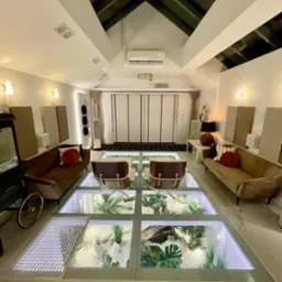 Salon z przeszkloną podłogą, pod którą znajduje się ogród. Nowoczesne wnętrze z sofami, krzesłami i wózkiem do popcornu. Widok na sufit z oknami.