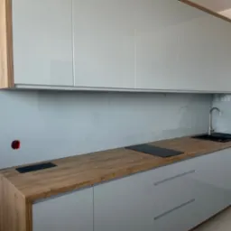 Nowoczesna kuchnia z szarymi, lakierowanymi szafkami, drewnianym blatem i jasnoniebieskim panelem szklanym. Widoczny zlew i płyta indukcyjna.