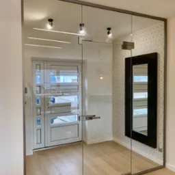 Nowoczesne, szklane drzwi wewnętrzne z metalową ramą i minimalistycznym uchwytem, widoczne oświetlenie sufitowe i geometryczna tapeta w tle. Jasne wnętrze.