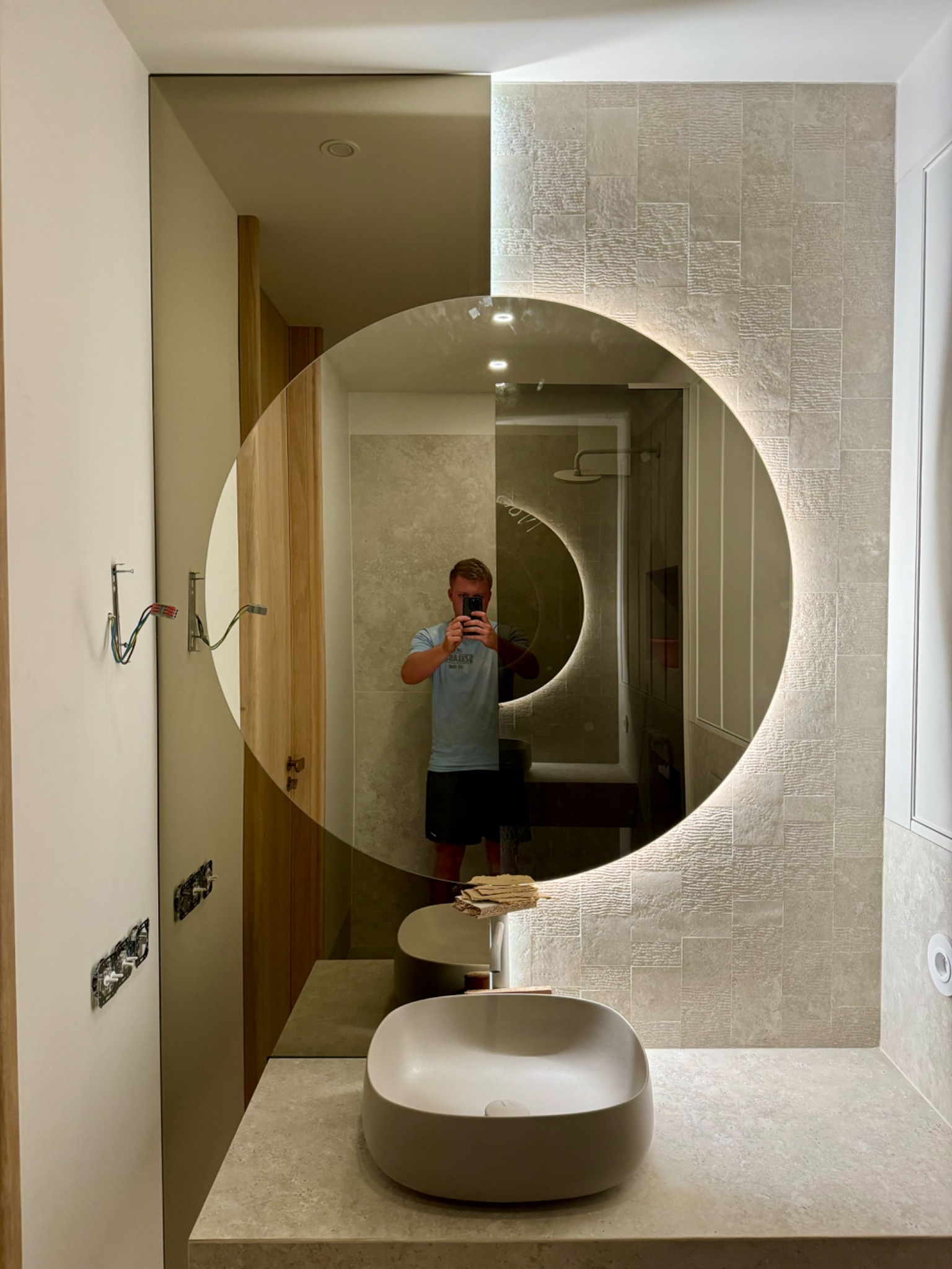 Łazienka z okrągłym lustrem LED. Widoczne płytki imitujące kamień, umywalka nablatowa i fragment instalacji elektrycznej. W lustrze odbicie osoby robiącej zdjęcie.