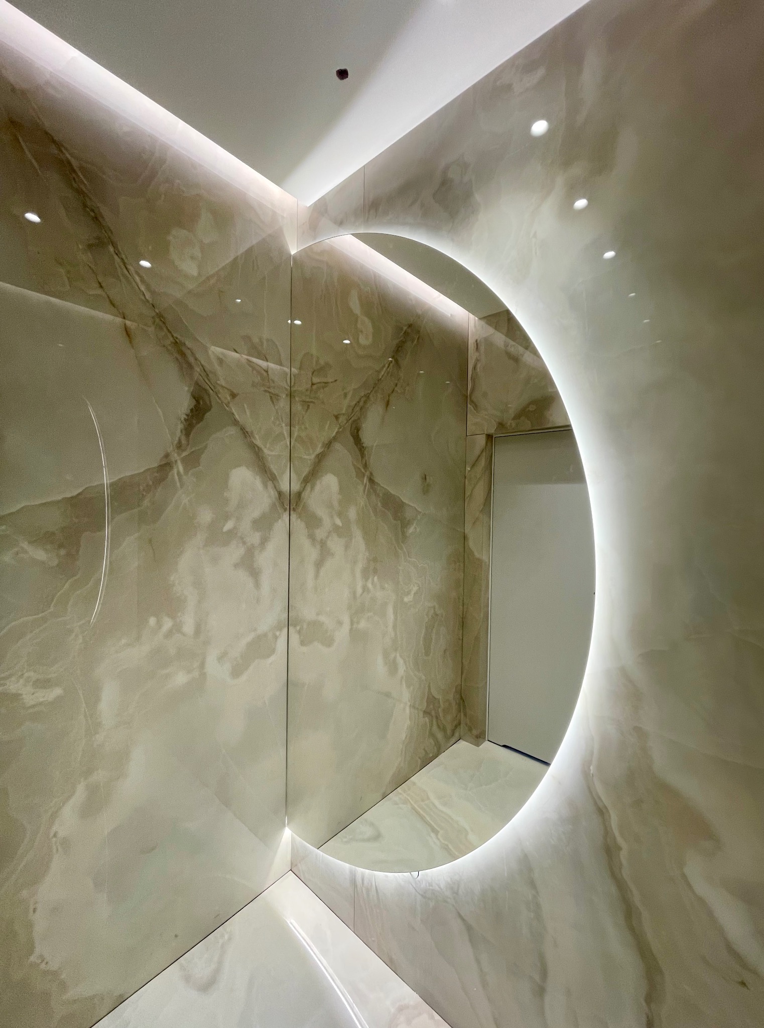 Nowoczesna łazienka z podświetlanym, okrągłym lustrem i marmurowymi płytkami na ścianach i podłodze. Widoczne delikatne oświetlenie LED na suficie.