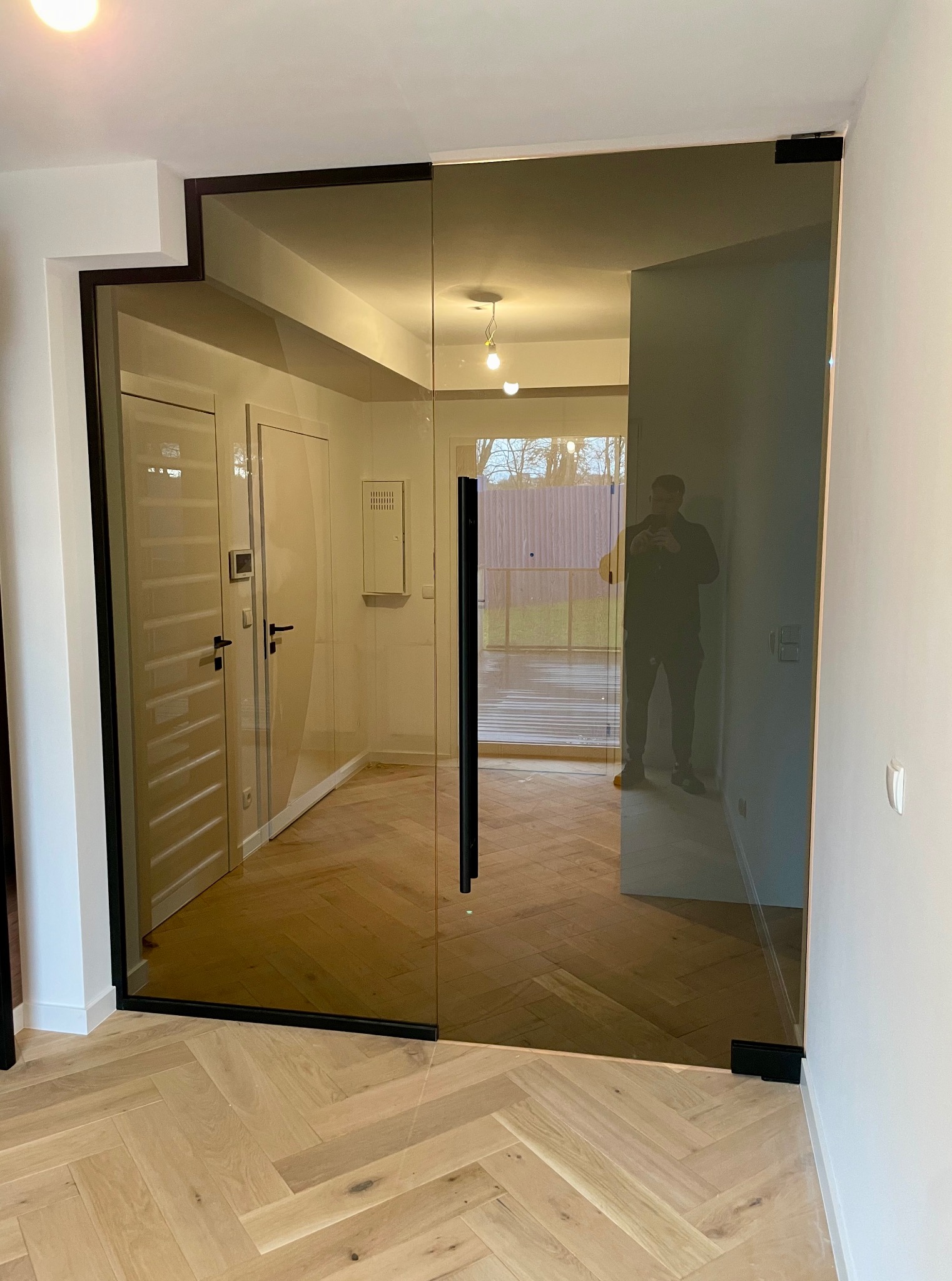 Szklane drzwi z czarną ramą w jasnym wnętrzu z parkietem w jodełkę. W tle widoczne inne drzwi i okno z widokiem na zewnątrz. Osoba widoczna w odbiciu.