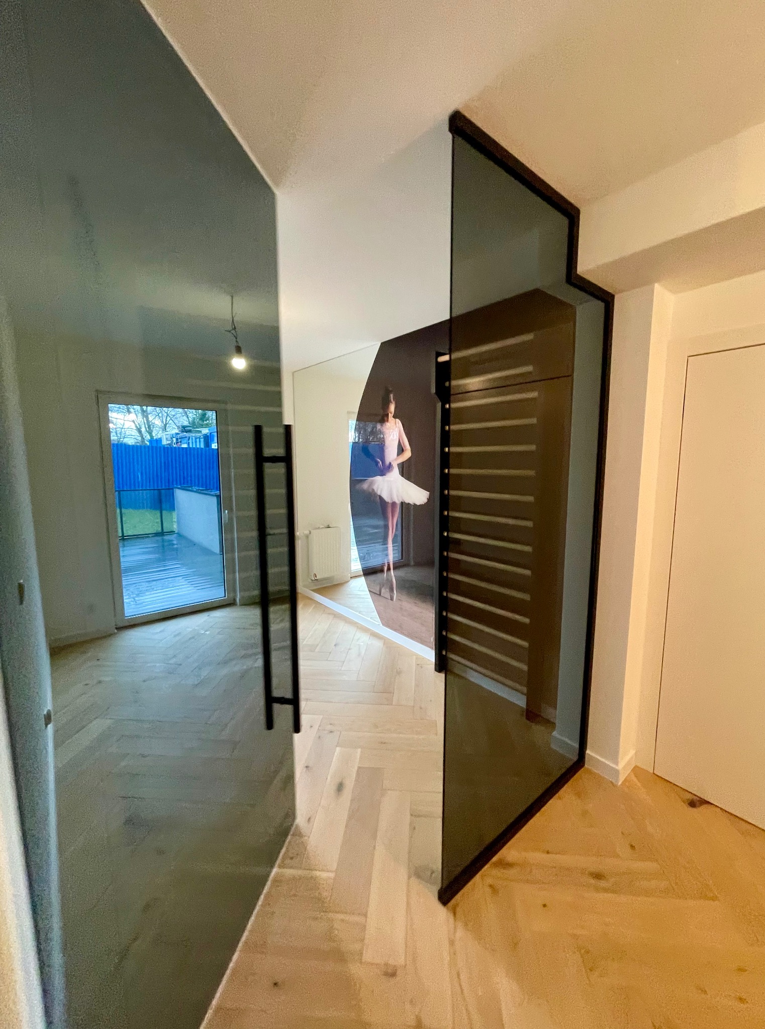 Szklane drzwi z czarną ramą, odbijające baletnicę, na tle jasnego parkietu i białych ścian. Nowoczesny design, elegancja i funkcjonalność.