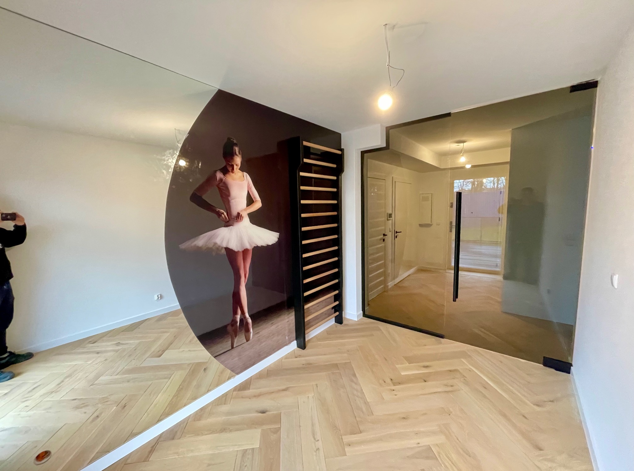 Nowoczesne wnętrze z lustrem z motywem baletnicy i szklanymi drzwiami z czarną ramą, parkiet w jodełkę. Widoczny fragment osoby robiącej zdjęcie.