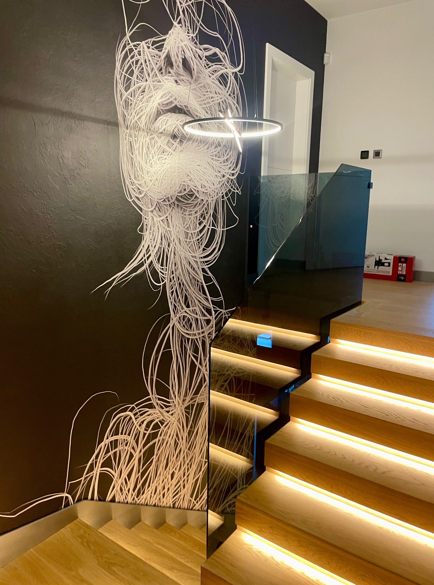 Nowoczesne schody z podświetleniem LED i szklaną balustradą. Na ścianie dekoracyjna grafika w stylu abstrakcyjnym. Minimalistyczne wnętrze z elementami drewna.