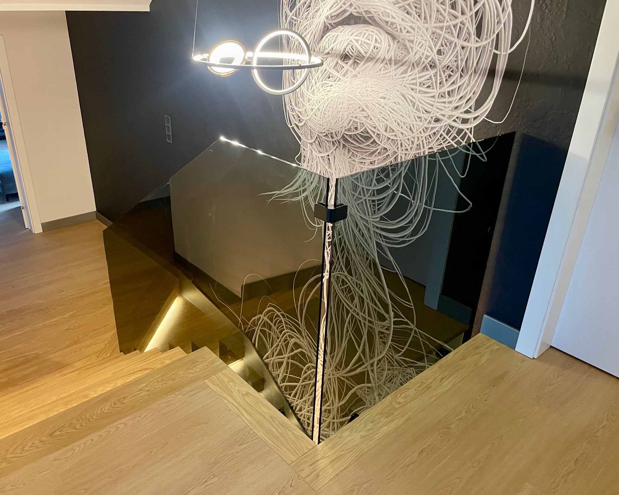 Nowoczesna, szklana balustrada schodowa z podświetleniem LED, odbijająca abstrakcyjny wzór na ścianie. Jasne, drewniane schody i minimalistyczne oświetlenie.