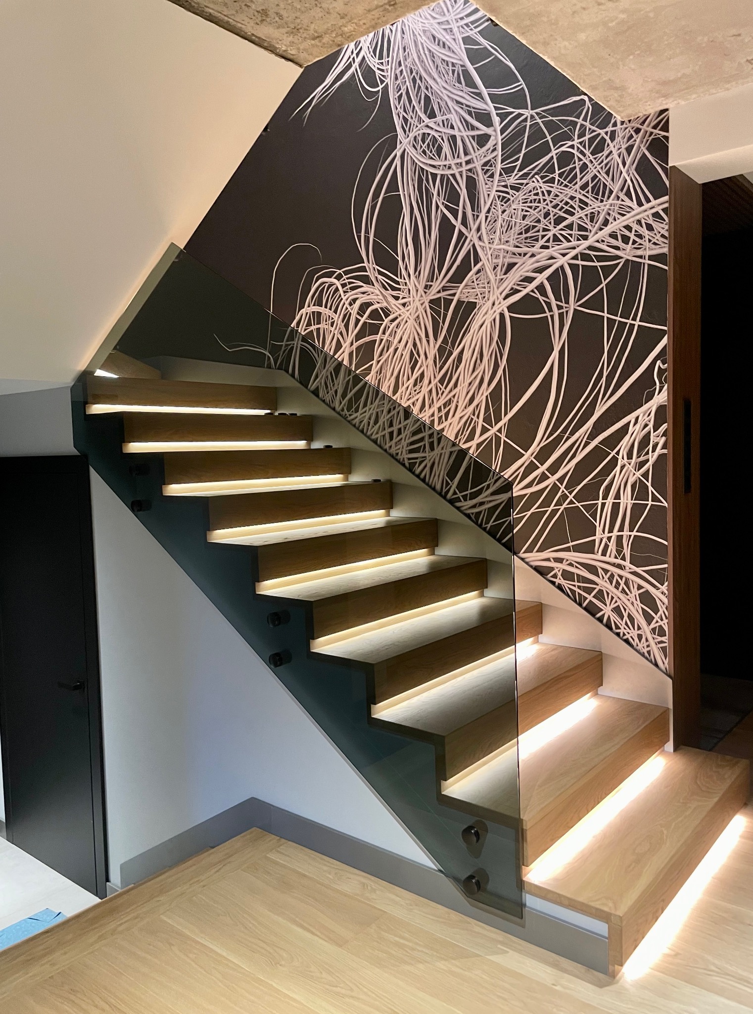 Drewniane schody z podświetleniem LED i szklaną balustradą, wspornikowe, z abstrakcyjnym wzorem na ścianie. Nowoczesne wykończenie wnętrza, jasne drewno.