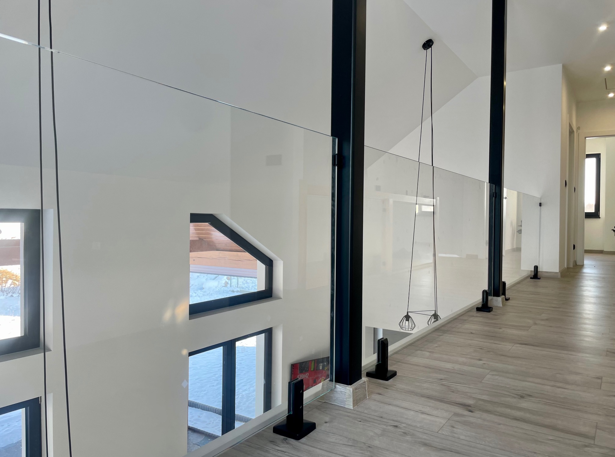 Nowoczesna balustrada ze szkła i czarnych słupków w jasnym wnętrzu. Widok z góry na drewnopodobną podłogę i okna z widokiem na zimowy krajobraz.