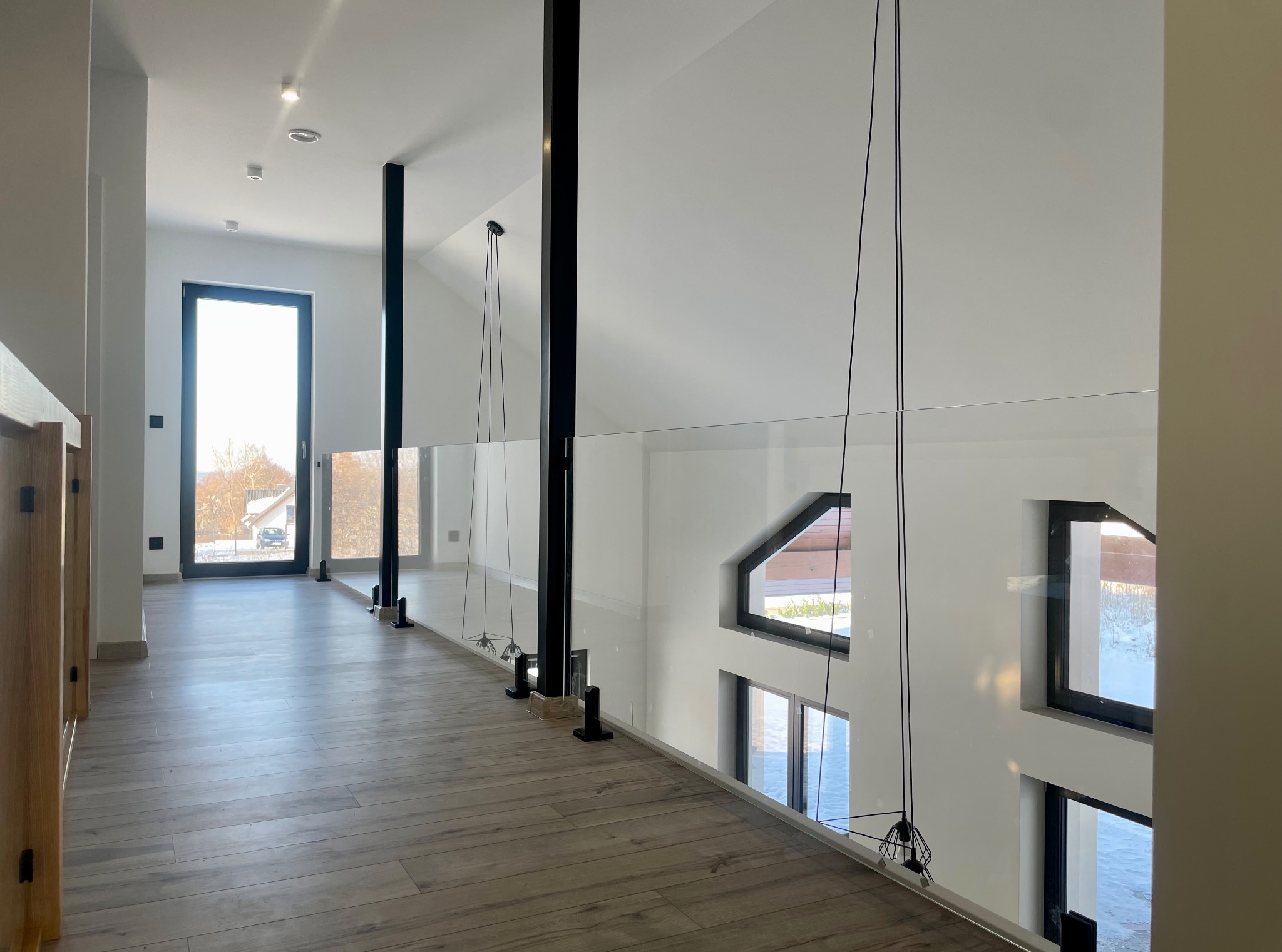 Nowoczesna balustrada szklana z czarnymi słupkami w jasnym wnętrzu domu. Widok na korytarz i okna. Podłoga z paneli imitujących drewno.