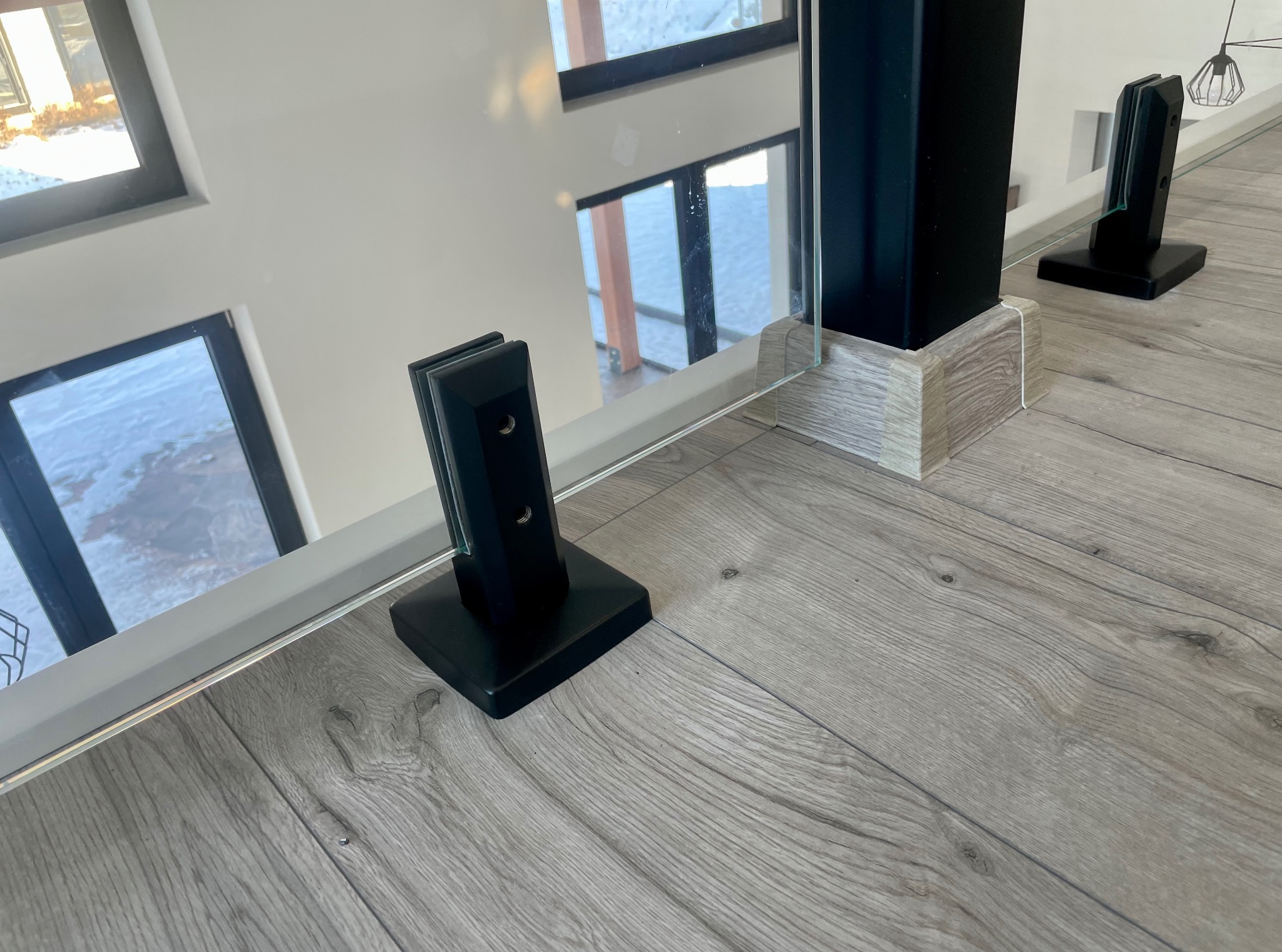 Szklana balustrada z czarnymi mocowaniami na podłodze imitującej drewno. Widok z góry na dół, okna w tle.