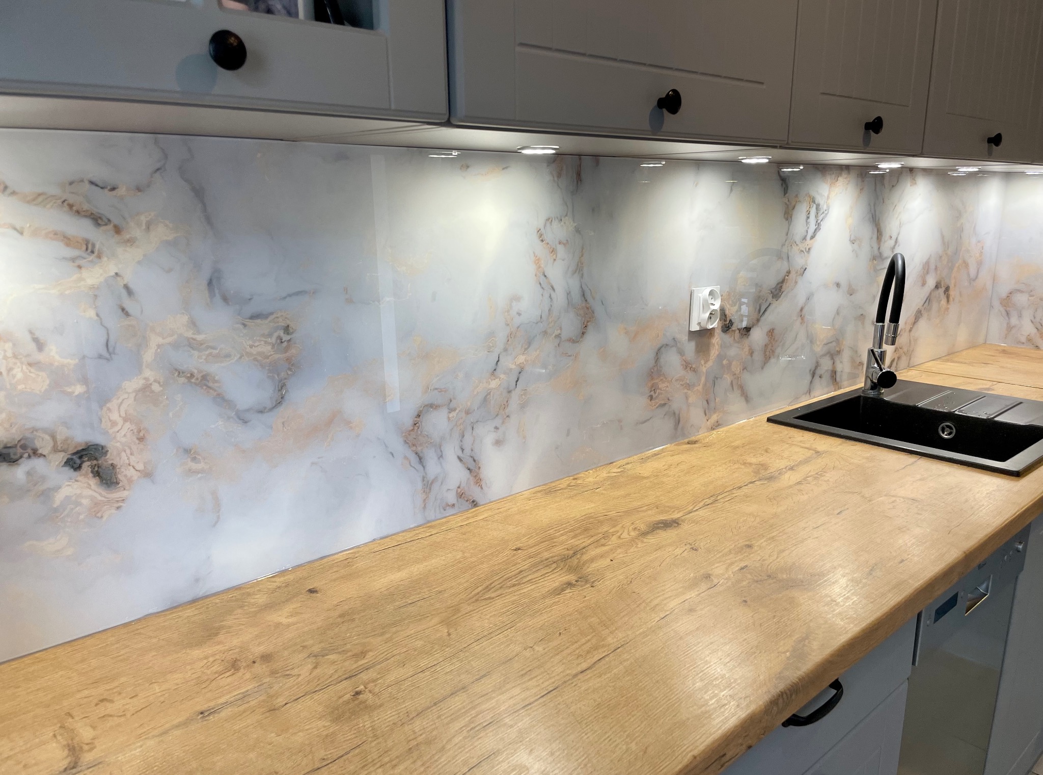 Szklany panel kuchenny z marmurkowym wzorem, oświetlony punktowo, zamontowany nad drewnianym blatem z czarnym zlewem i baterią. Nowoczesna aranżacja kuchni.