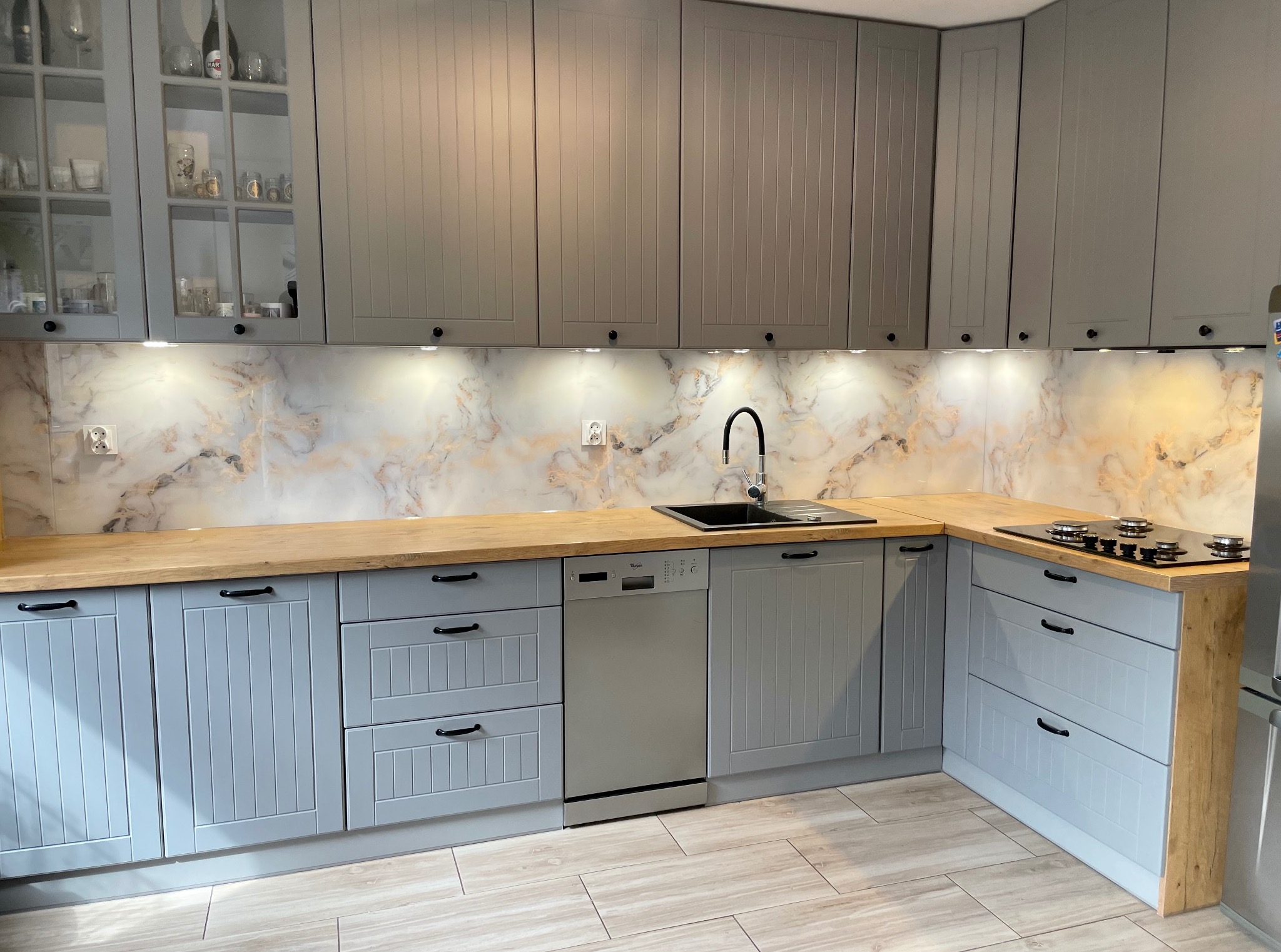 Jasna kuchnia w stylu rustykalnym z drewnianym blatem i marmurowym backsplashiem. Szafki górne szare, dolne błękitne. Widoczny zlew, płyta grzewcza i sprzęt AGD.
