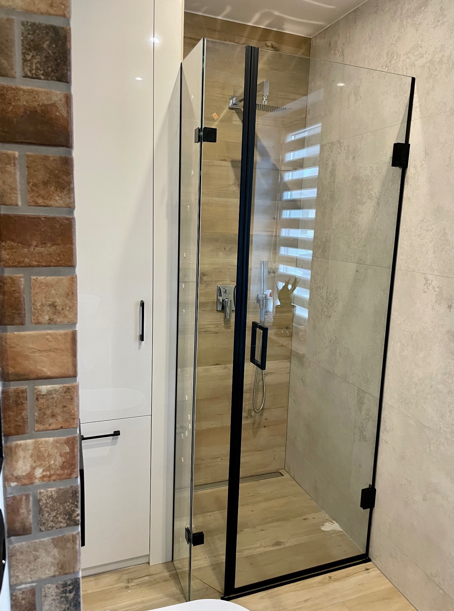 Kabina prysznicowa ze szklanymi drzwiami, czarne okucia, drewnopodobne płytki wewnątrz, obok biała szafka i fragment ściany z cegły. Nowoczesna łazienka.