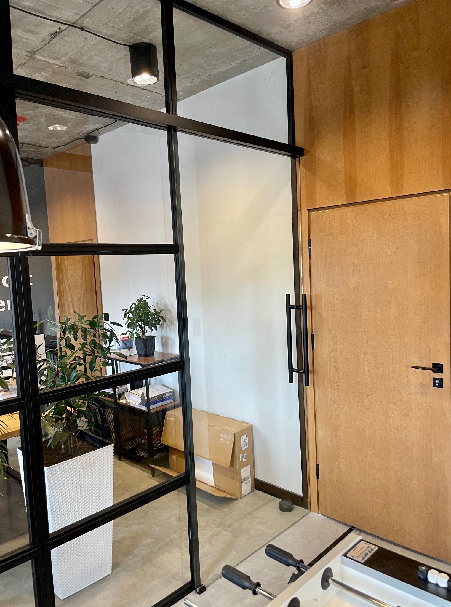 Nowoczesne biuro z przeszklonymi ścianami w czarnych ramach i drewnianymi drzwiami. Widoczne rośliny doniczkowe i fragment stołu do piłkarzyków.