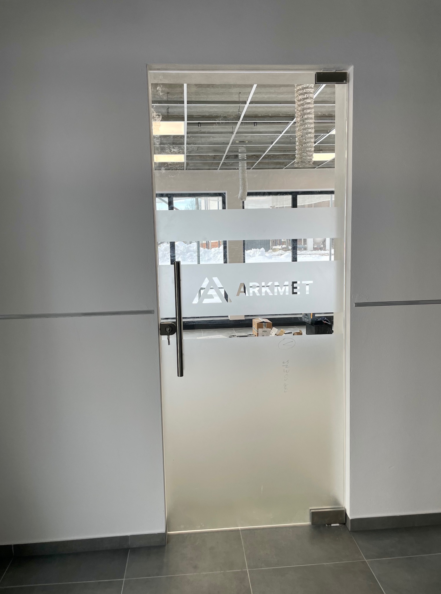Szklane drzwi z matową folią i logo firmy Arkmet, widoczne przez szybę wnętrze w trakcie prac wykończeniowych, minimalistyczny design w szarościach.