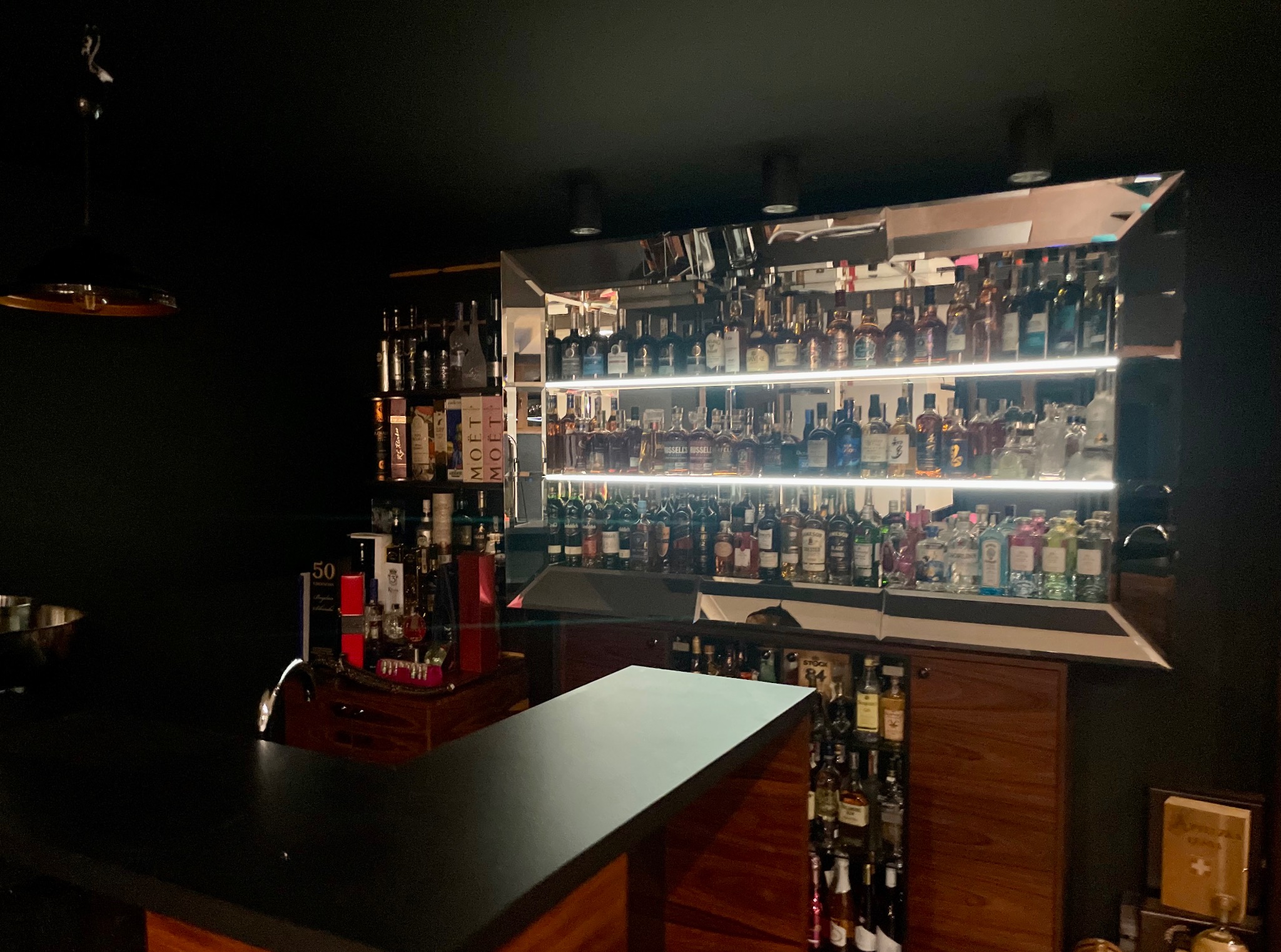 Domowy bar z oświetloną, lustrzaną ścianą za blatem. Butelki alkoholi ustawione na półkach. Ciemne ściany, ciepłe oświetlenie. Nowoczesny design.